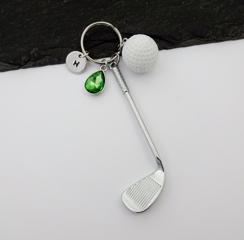 Puede incluir: Un llavero de palo de golf plateado con una bola de golf blanca, un amuleto de cristal verde y un amuleto plateado con la letra "H" en &eacute;l.