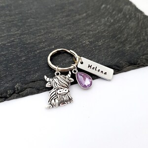 Puede incluir: Un llavero plateado con un dije de vaca de las Tierras Altas, una gema morada en forma de l&aacute;grima y una etiqueta rectangular con el nombre "Helena" grabado. El llavero est&aacute; en un anillo plateado, sobre un fondo gris oscuro.