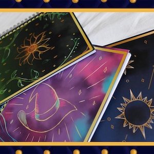 Puede incluir: Tres cuadernos de espiral con tapas negras y diseños dorados. El primer cuaderno tiene un diseño floral verde y dorado con un girasol en el centro. El segundo cuaderno tiene un diseño morado y dorado con una explosión de estrellas y un sombrero de bruja. El tercer cuaderno tiene un diseño negro y dorado con un sol y estrellas.