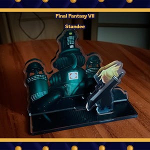 Könnte beinhalten: Ein Aufsteller mit einer Szene aus Final Fantasy VII. Der Aufsteller zeigt eine dunkelgrüne Industriestruktur und eine Figur mit einem Schwert. Der Text "Final Fantasy VII Standee" ist oben sichtbar.