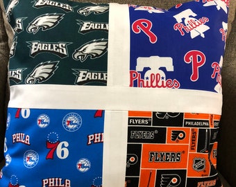 May include: A decorative pillow featuring four sections with sports team logos. The sections display logos for the Philadelphia Eagles, Phillies, 76ers, and Flyers. The pillow has a white border and is placed on a gray surface.