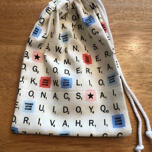 May include: A drawstring bag with a cream-colored background, patterned with Scrabble tiles. The bag features a white drawstring closure. The tiles include letters, numbers, and special squares like "Triple Letter Score".