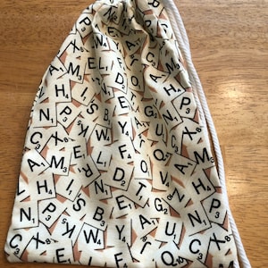 May include: A white drawstring bag with a pattern of brown and white Scrabble tiles. The tiles have letters and numbers on them.