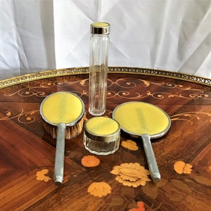 May include: A vintage yellow and silver vanity set with a brush, two mirrors, a jar, and a bottle. The set is on a wooden table with a floral design.