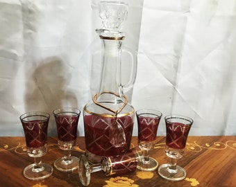 May include: A set of five clear glass shot glasses with a red and gold design. The glasses are arranged in a semi-circle around a clear glass decanter with a red and gold design.
