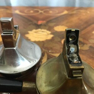 May include: Two vintage table lighters, one silver and one brass. The silver lighter has a round, domed top and a black handle. The brass lighter has a round, domed top and a black flint wheel.