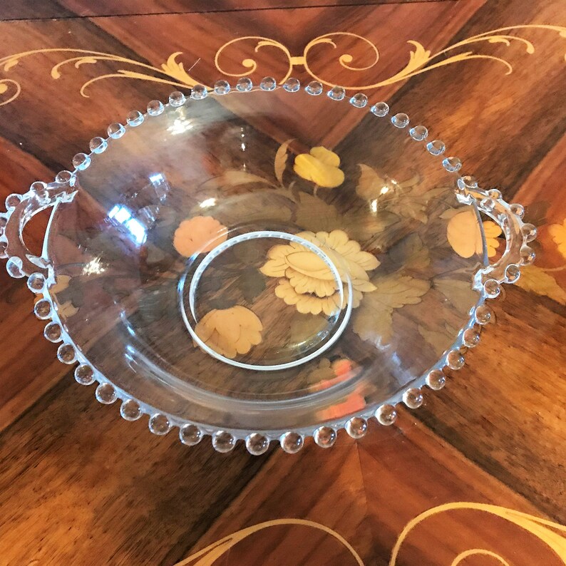 K&ouml;nnte beinhalten: Eine klare Glassch&uuml;ssel mit einem perlenbesetzten Rand und zwei Griffen. Die Sch&uuml;ssel steht auf einer Holzoberfl&auml;che mit einem Blumendesign.