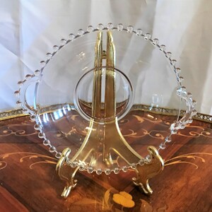 K&ouml;nnte beinhalten: Eine klare Glassch&uuml;ssel mit einem perlenbesetzten Rand und zwei Griffen. Die Sch&uuml;ssel steht auf einem goldenen Metallst&auml;nder.