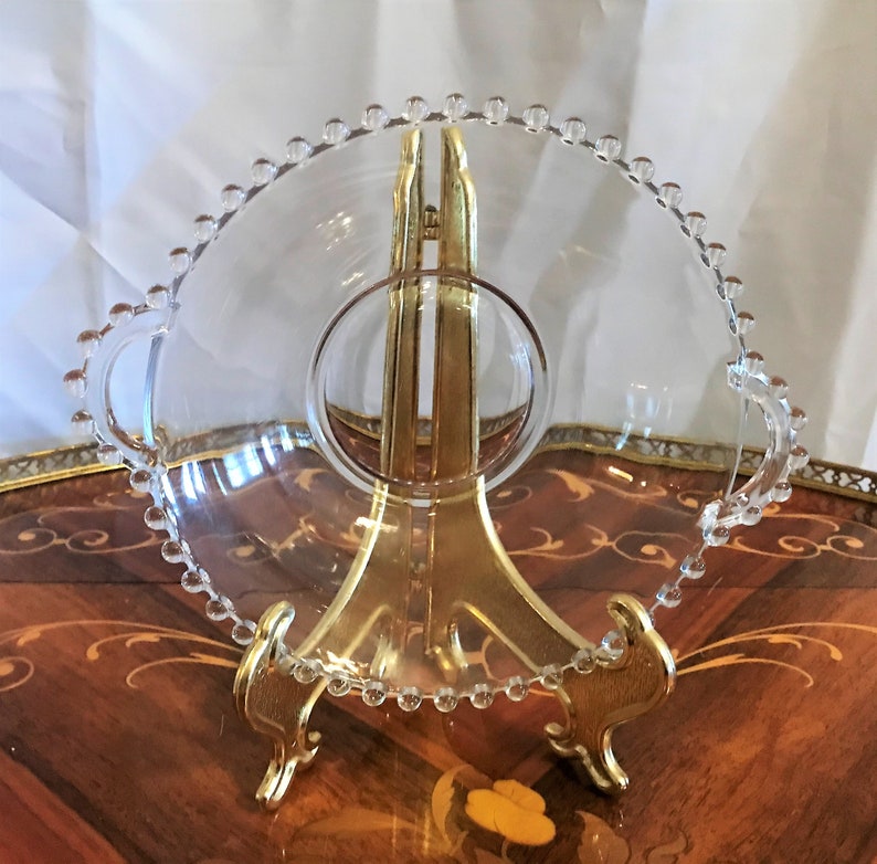 K&ouml;nnte beinhalten: Eine klare Glassch&uuml;ssel mit einem perlenbesetzten Rand und zwei Griffen. Die Sch&uuml;ssel steht auf einem goldenen Metallst&auml;nder.