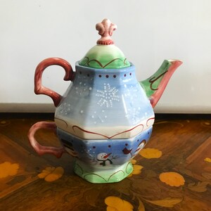 May include: A decorative teapot with a matching teacup base. The teapot is painted with a winter theme, featuring snowflakes, snowmen, and a red bird. The handle and spout have red and green accents. The lid is topped with a pink and green finial.