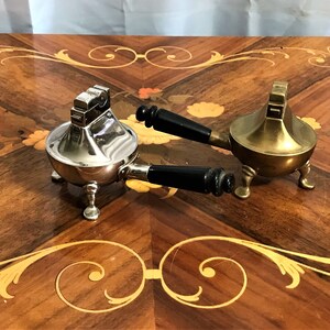May include: Two vintage table lighters with black handles. One is silver and the other is brass. Both have three legs and are on a wooden table.