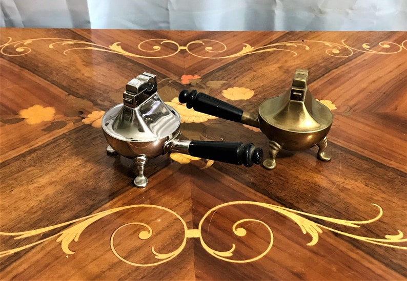 May include: Two vintage table lighters with black handles. One is silver and the other is brass. Both have three legs and are on a wooden table.