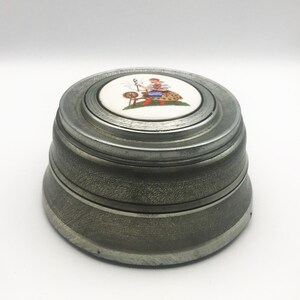 May include: A round, metallic lidded container with a brushed gray finish. The center of the lid features a circular white ceramic inlay decorated with a colorful illustration of a person sitting at a spinning wheel on a patch of green grass.