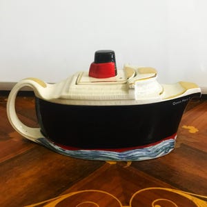 May include: A ceramic teapot shaped like a ship, featuring a black hull, cream-colored superstructure, and a red and black smokestack. The teapot has a handle and spout, with the words "Queen Mary 2" on the side. The base has a blue and red wave design.
