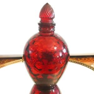 May include: A ruby red glass bottle with a decorative stopper. The bottle has a rounded body and a textured surface. The stopper is a spiral design. The bottle is likely for perfume or other liquids.