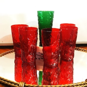 May include: A set of six textured glass tumblers, five in a vibrant red and one in green. The glasses are arranged on a mirrored tray with a gold rope trim. The tumblers have a unique, organic pattern, reflecting light and adding visual interest.