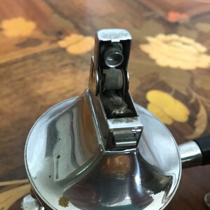 May include: A silver-toned table lighter with a round base and a rectangular top. The lighter has a silver-toned knob on the top and a black handle.