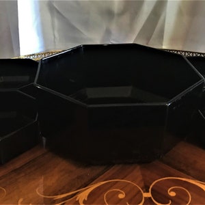 Pode incluir: Um conjunto de pratos de servir octogonais pretos, com um prato central maior e quatro pratos menores ao redor. Os pratos est&atilde;o dispostos sobre uma superf&iacute;cie de madeira com um padr&atilde;o decorativo.