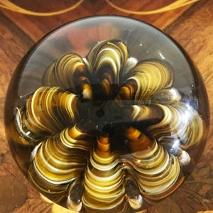 May include: A clear glass paperweight with an internal design of swirling brown, white, and yellow glass. The paperweight has a rounded shape and a smooth surface. The design resembles a flower or abstract form.