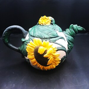 May include: A ceramic teapot featuring a large, raised yellow sunflower with a dark brown center on the side. The lid is adorned with green leaves and a small yellow flower. The handle and spout are sculpted to resemble dark green stems.