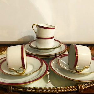 May include: A set of white porcelain teacups and saucers with a burgundy and gold trim. The teacups have a curved handle and are stacked on matching saucers. The set is arranged on a gold-rimmed serving tray, perfect for a tea party.