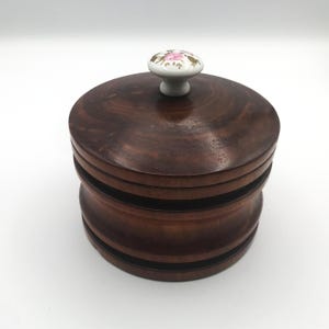 May include: A round, dark brown wooden container with a lid. The lid features a small, white ceramic knob decorated with pink and gold floral details. The container has a stacked design with visible wood grain.