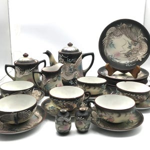 May include: A tea set featuring a teapot, sugar bowl, creamer, cups, saucers, plates, and two small shakers. Each piece is crafted from dark brown and cream ceramic with raised, detailed dragon motifs and subtle pink and blue accents.