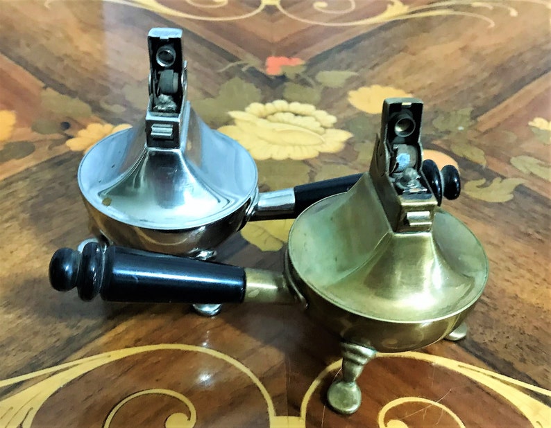 May include: Two vintage table lighters with black handles. One is silver and the other is brass.