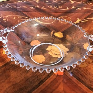 K&ouml;nnte beinhalten: Eine klare Glassch&uuml;ssel mit perlenbesetzten R&auml;ndern und zwei Griffen. Die Sch&uuml;ssel ist leer und steht auf einer Holzoberfl&auml;che.