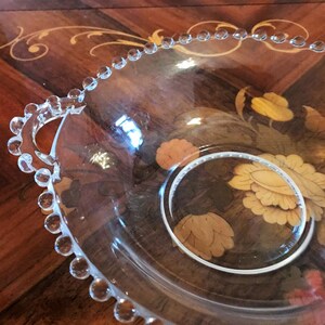 K&ouml;nnte beinhalten: Eine klare Glassch&uuml;ssel mit einem perlenbesetzten Rand und zwei Griffen. Die Sch&uuml;ssel steht auf einer Holzoberfl&auml;che mit einem Blumendesign.