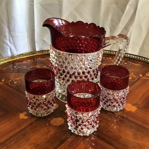 May include: A set of four clear glass drinking glasses with a red and white textured design. The glasses are arranged on a wooden table with a pitcher in the center.