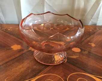May include: A pink glass bowl with a scalloped rim and a footed base. The bowl has a delicate etched floral design.