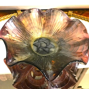 May include: An iridescent glass bowl with a ruffled rim and a star-shaped design in the center. The bowl has a dark purple and gold color scheme, with a textured, radiating pattern. The bowl is likely for decorative purposes.