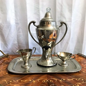 May include: A silver serving set with a coffee pot, creamer, and sugar bowl on a decorative tray. The coffee pot has a spout and a handle on each side. The creamer and sugar bowl have handles on one side.