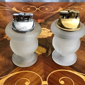 May include: Two frosted glass vintage lighters with metal tops. One lighter has a silver top and the other has a gold top. The lighters are sitting on a wooden surface with a decorative inlay.