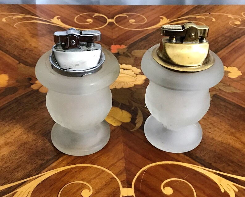May include: Two frosted glass vintage lighters with metal tops. One lighter has a silver top and the other has a gold top. The lighters are sitting on a wooden surface with a decorative inlay.
