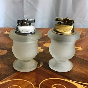 May include: Two frosted glass table lighters with silver and gold tops. The lighters are shaped like urns with a round base.
