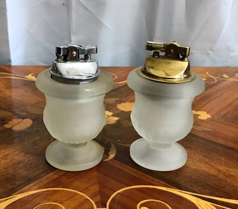 May include: Two frosted glass table lighters with silver and gold tops. The lighters are shaped like urns with a round base.