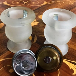May include: Two frosted glass candle holders with a brass lid on a wooden table. The candle holders are in the shape of urns with a round base.
