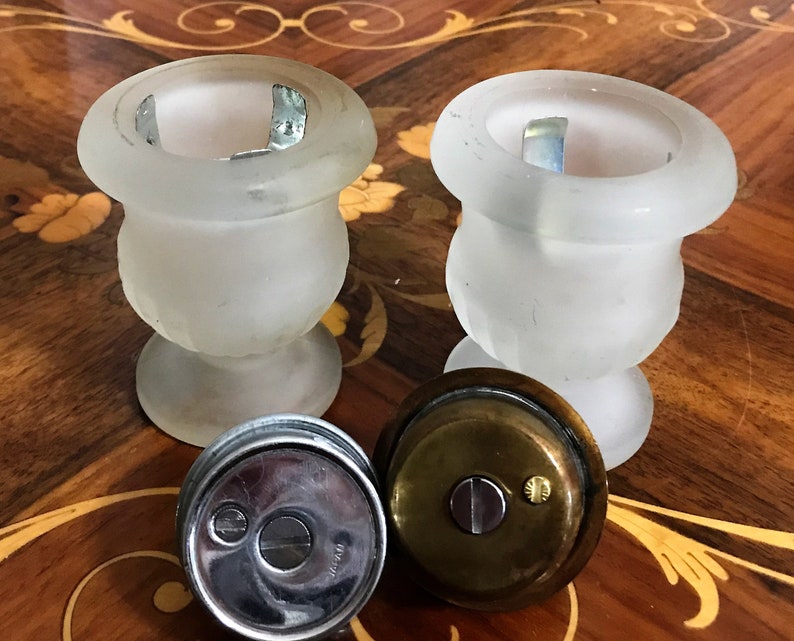 May include: Two frosted glass candle holders with a brass lid on a wooden table. The candle holders are in the shape of urns with a round base.