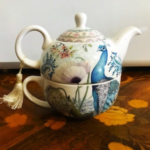 May include: A white ceramic tea-for-one set featuring a peacock design. The teapot and teacup are adorned with blue peacock illustrations, floral patterns, and a tassel. The set is designed for a single serving of tea.