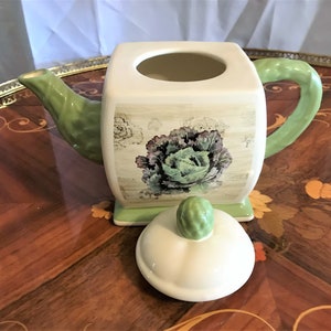 May include: A square ceramic teapot with a green lid and handle. The teapot has a floral design with a cabbage in the center. The teapot is sitting on a wooden table with a matching lid next to it.