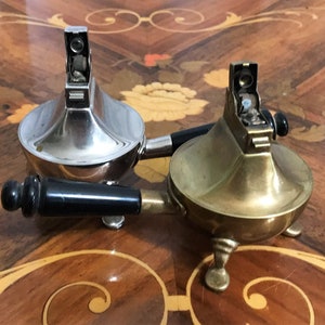May include: Two vintage table lighters, one silver and one brass, with black handles. The lighters have a distinctive design with a round base and a tall, slender neck.
