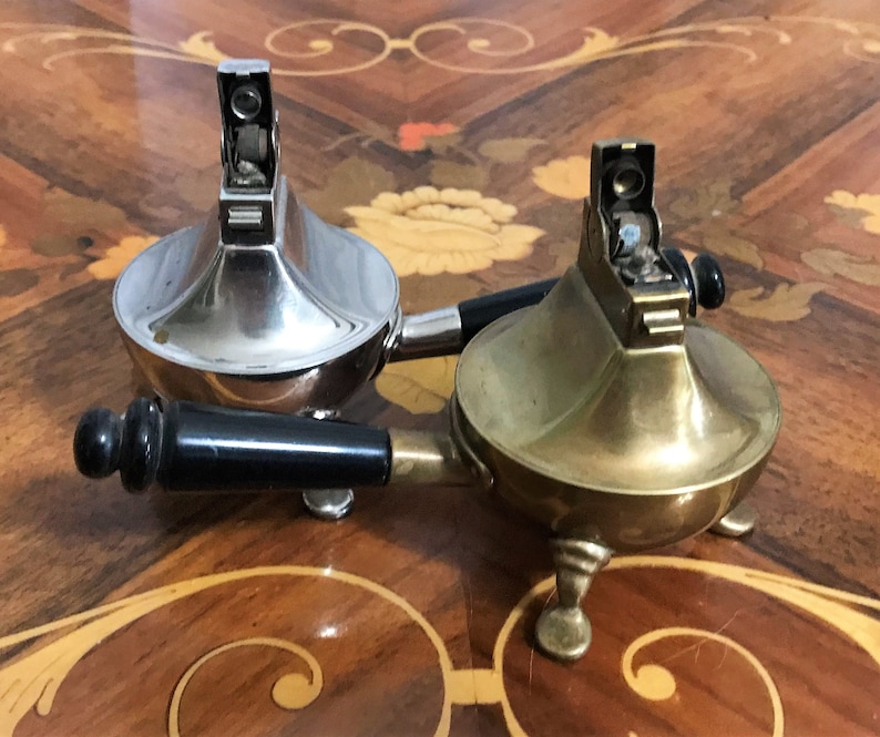 May include: Two vintage table lighters, one silver and one brass, with black handles. The lighters have a distinctive design with a round base and a tall, slender neck.
