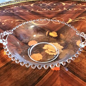K&ouml;nnte beinhalten: Eine klare Glassch&uuml;ssel mit perlenbesetzten R&auml;ndern und zwei Griffen. Die Sch&uuml;ssel steht auf einer Holzoberfl&auml;che mit einem Blumenmuster.