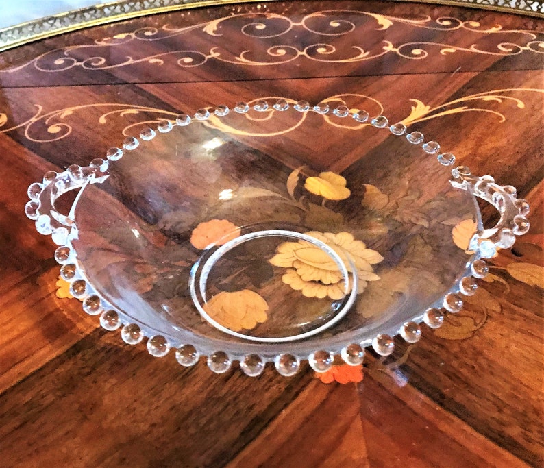 K&ouml;nnte beinhalten: Eine klare Glassch&uuml;ssel mit perlenbesetzten R&auml;ndern und zwei Griffen. Die Sch&uuml;ssel steht auf einer Holzoberfl&auml;che mit einem Blumenmuster.