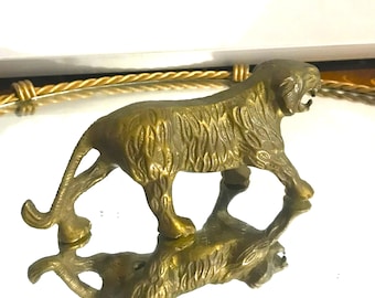 May include: A brass tiger figurine with detailed fur texture, posed in a walking stance. The figurine is reflected on a shiny surface, creating a mirrored image. The tiger's mouth is open, and its tail is raised.
