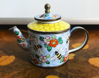 Puede incluir: Pequeña tetera decorativa con cuerpo azul claro adornado con abejas y motivos florales. La tapa es amarilla con diseño de panal, y el asa y el pico tienen detalles dorados. Una pieza decorativa encantadora.