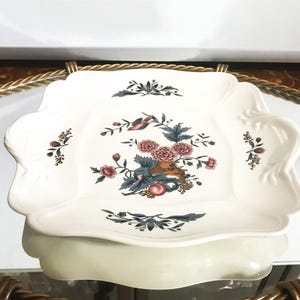 May include: A white, scalloped-edge serving dish with a floral and bird design. The center features a bouquet of pink flowers, green leaves, and a bird. The dish is likely made of ceramic or porcelain.
