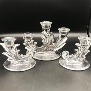 May include: A set of three clear glass candle holders featuring a decorative scrollwork design. The central piece holds three candles, while the two flanking pieces are single candle holders, all resting on circular bases with molded leaf patterns.